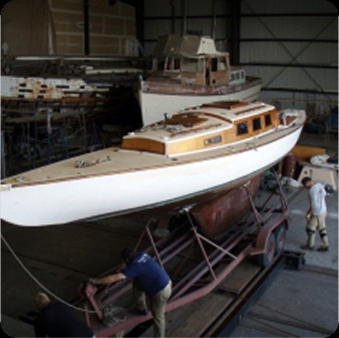 Three people work together to move a sleek wooden sailboat, one of the classic Square Metre Yachts, on a trailer inside a large boatyard or warehouse, with other boats visible in the background.