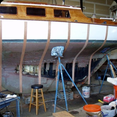 A wooden boat, reminiscent of classic Square Metre Yachts, is being repaired in a garage with vertical wooden ribs installed along its hull. Tools, a stool, paint tins, and support stands are scattered around the workspace.