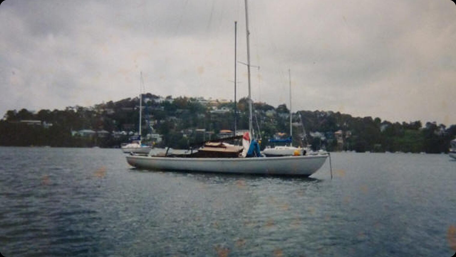 A white Schärenkreuzer is anchored on calm water with a hilly, tree-lined shoreline and houses in the background under a cloudy sky.