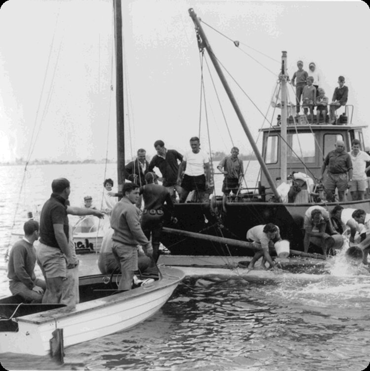 Black and white photo of people on two Classic Sailing Yachts. Several individuals are gathered at the edge, appearing to work or pull something from the water, whilst others observe or relax on the deck and roof.