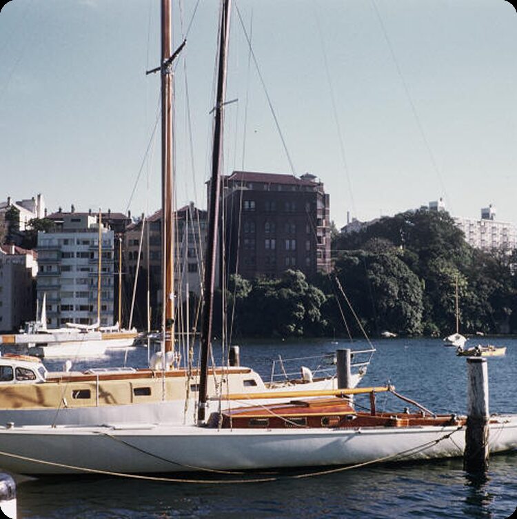 A Schärenkreuzer sailboat moored in a marina with blocks of flats and trees visible in the background, under a clear blue sky.