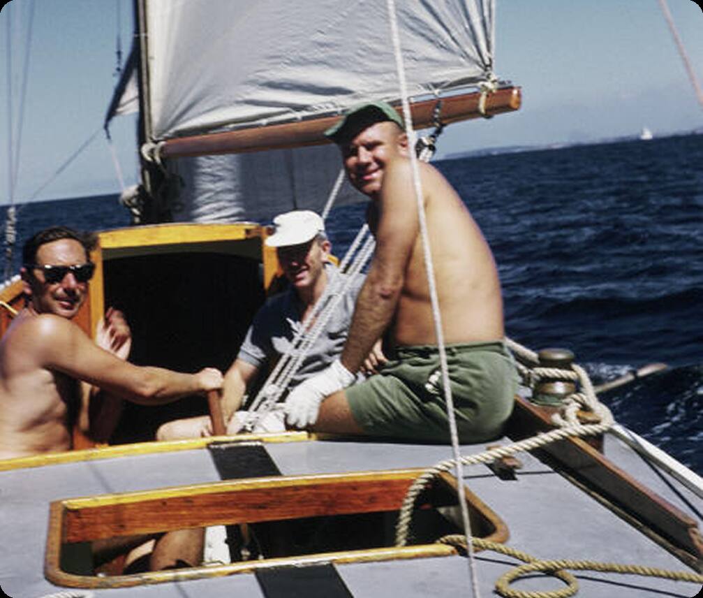 Three men sit and smile on the deck of a Classic Sailing Yacht on a sunny day, with deep blue sea and clear sky in the background. One man is bare-chested and wearing a green cap, while the others wear hats and sunglasses.