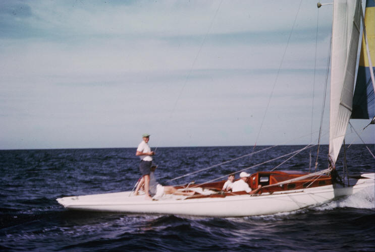 Three people are sailing on a sleek white Skerry Cruiser under a clear blue sky. One stands near the bow, while two relax beneath the cabin’s wooden structure. The calm water and classic Schärenkreuzer lines complete this serene scene.