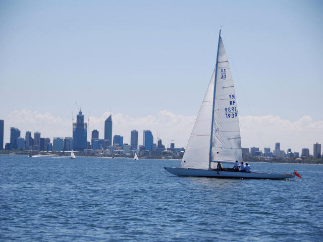 A white sailboat, reminiscent of classic sailing yachts, glides on calm blue water as passengers enjoy views of a city skyline and high-rise buildings under a clear sky.