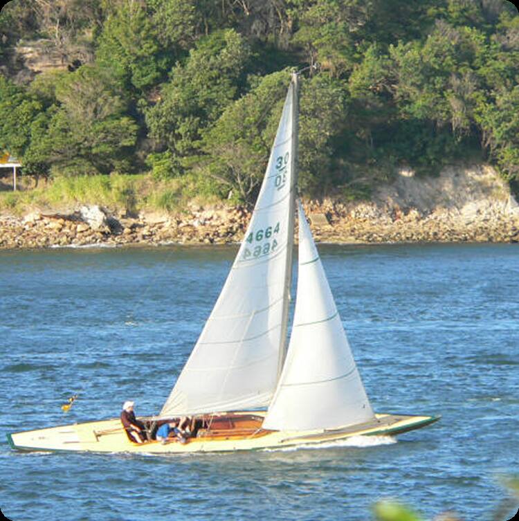 A small Skerry Cruiser with two people on board sails on a calm blue river, with a rocky and tree-lined shore in the background under a sunny sky.