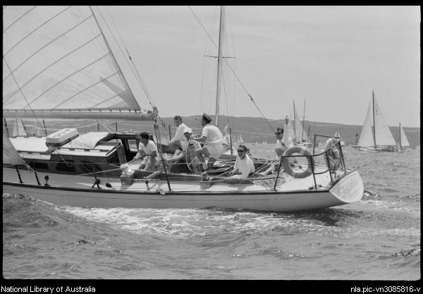 A group of people in casual clothing sail a classic sailing yacht on the sea, working together with the sails and ropes. Other Square Metre Yachts are visible in the background under a partly cloudy sky.