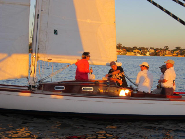 A group of people sit and stand on a classic sailing yacht named Pachani on calm water during sunset, with buildings and trees visible in the background.