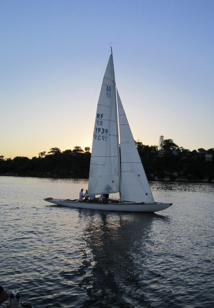 A white Skerry Cruiser with three people aboard glides on calm water at sunset, its tall sail marked with numbers and letters. Trees and a dark shoreline frame this classic sailing yacht beneath the softly lit, clear sky.