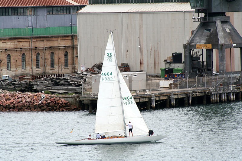 A white Skerry Cruiser sailboat with four people aboard sails near an industrial waterfront with warehouses, a crane, stacked timber, and rocks along the shore. The tall sails are marked with numbers and letters.