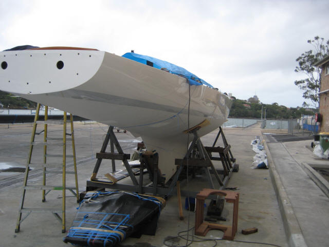 A white Classic Sailing Yacht is propped up on wooden stands in a boatyard, partially covered with a blue tarpaulin. Ladders, tools, and equipment are scattered around on the concrete ground under a cloudy sky.