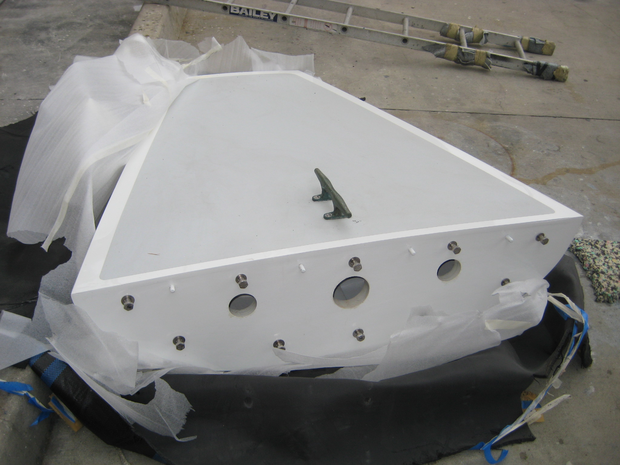 A large, white, triangular metal panel with several circular holes and a green handle—likely a part of Classic Sailing Yachts—rests on protective padding on a concrete floor. A ladder lies in the background.