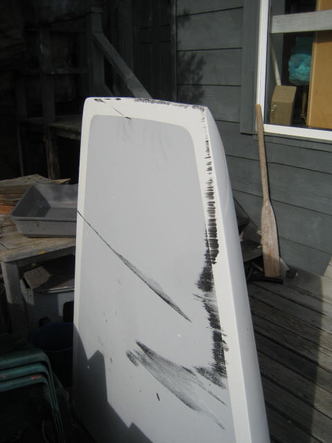 A large white object with black scuff marks, reminiscent of a Schärenkreuzer hull, leans against a grey wooden wall on a deck. A wooden paddle and various tools are nearby, with sunlight casting shadows across the scene.