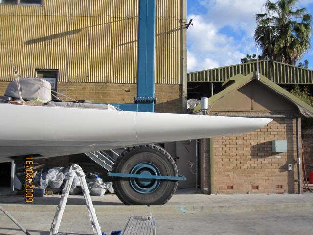 A close-up side view of a white Square Metre Yachts’ pointed bow on a trailer, with a large wheel beneath. Industrial buildings and a palm tree are visible in the background. The photo is dated 18/09/2009.