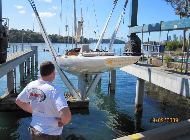 A man stands on a dock, watching a white Skerry Cruiser sailboat being lifted by a crane over the water. Trees, a bridge, and city buildings are visible in the background. The date on the photo is 19/09/2009.