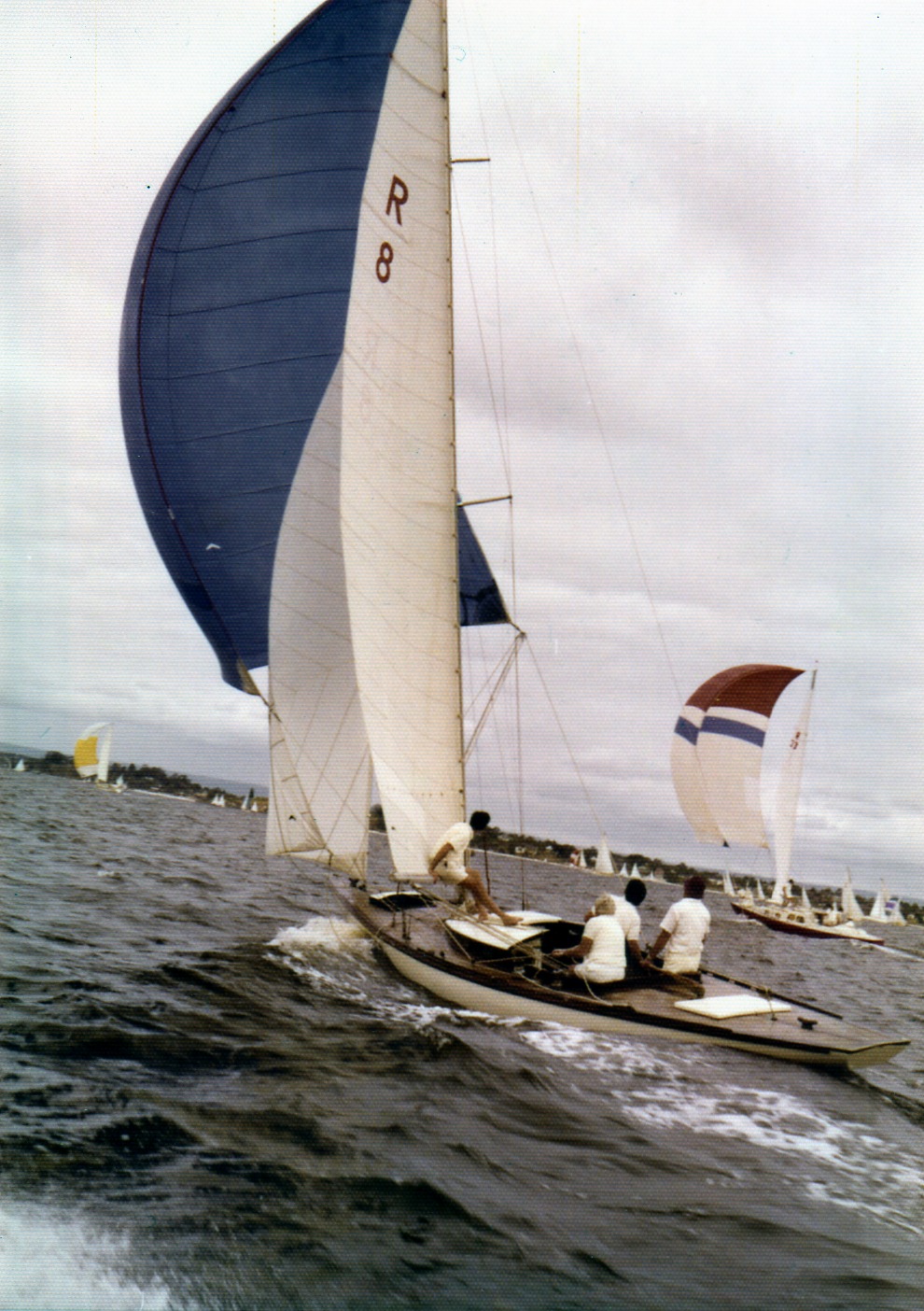 A sailboat with a large blue and white sail glides across the water, crewed by three people. Other Classic Sailing Yachts are visible in the background under a cloudy sky as waves create splashes along the boat’s side.