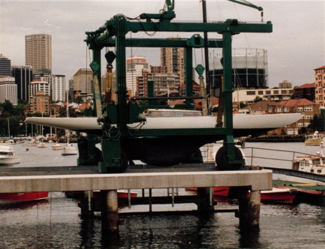 A classic sailing yacht is suspended on a green boat lift above a dock, with city buildings and moored boats visible in the background beside the waterfront.