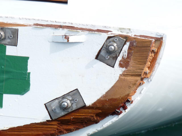 Close-up of a damaged white boat hull, possibly from a Classic Sailing Yacht or Square Metre Yacht, with exposed wood and chipped paint, reinforced by metal plates and bolts. Green tape on the left side suggests ongoing repairs.