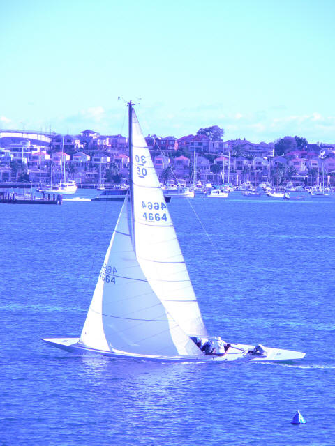 A Classic Sailing Yacht with three people aboard glides on a calm blue body of water, with houses, trees, and other boats visible along the shore in the background under a clear sky.