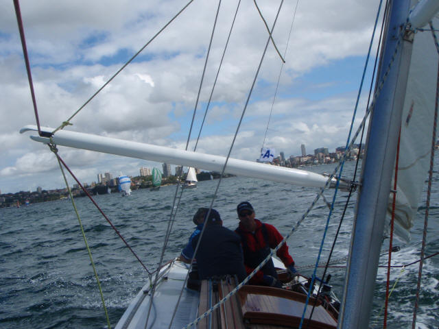 Two people are sailing a Square Metre Yacht on choppy water under a cloudy sky, with several other sailboats and a city skyline visible in the background.