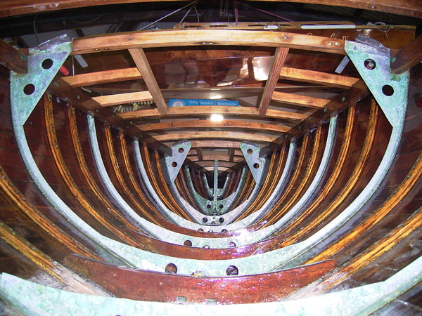 Interior view of a wooden Schärenkreuzer hull under construction, showing curved ribs, metal supports, and wooden framework. The photo from inside towards the bow highlights craftsmanship and structural details of classic sailing yachts.