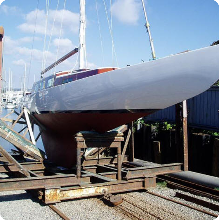 A white sailboat with a red underside sits on a wooden and metal cradle by the water, ready for launch. Blue sky and other Classic Sailing Yachts are visible in the background.
