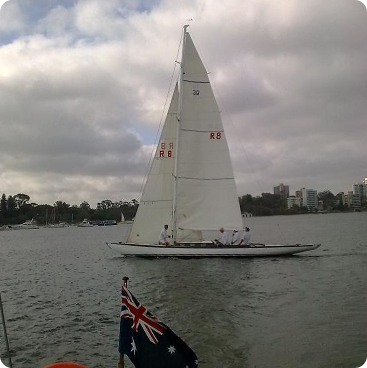 A white Square Metre Yacht with three people onboard sails on a calm body of water under a cloudy sky. An Australian flag is visible in the foreground, with trees and buildings lining the shore in the background.