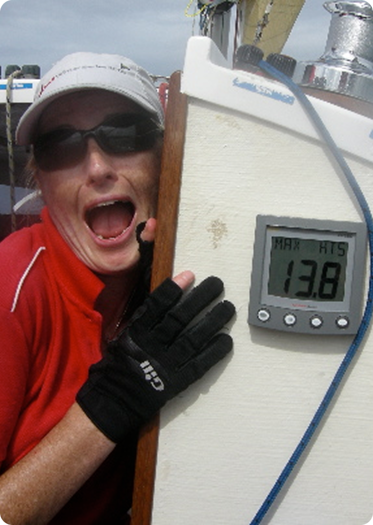A person in sunglasses and a red shirt excitedly poses next to a digital gauge on a Classic Sailing Yacht showing MAX 13.8. The person is smiling widely, wearing black gloves and a white cap.