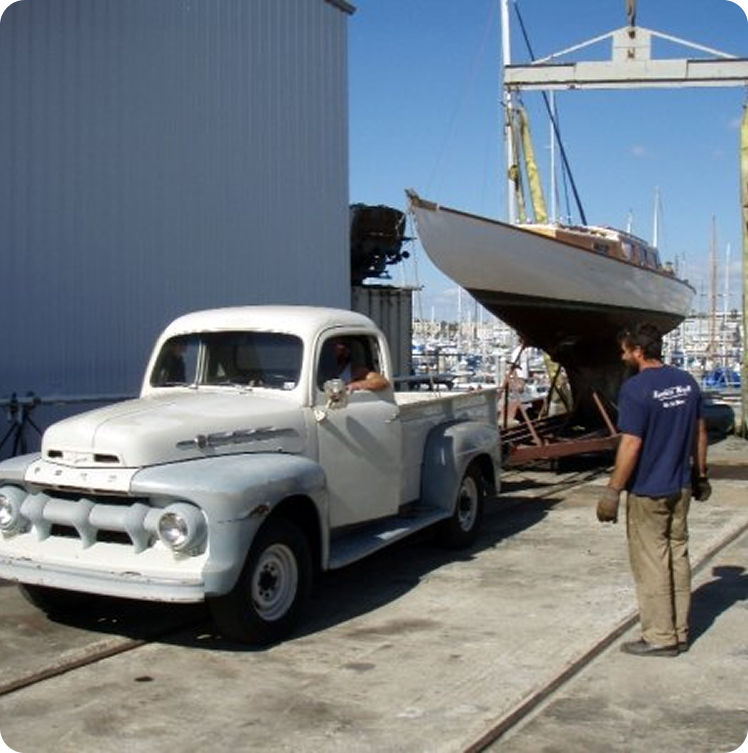 A vintage white pickup truck is parked near a boatyard, where a large Skerry Cruiser sailboat is being lifted by a crane; a man in work clothes stands nearby watching the scene.