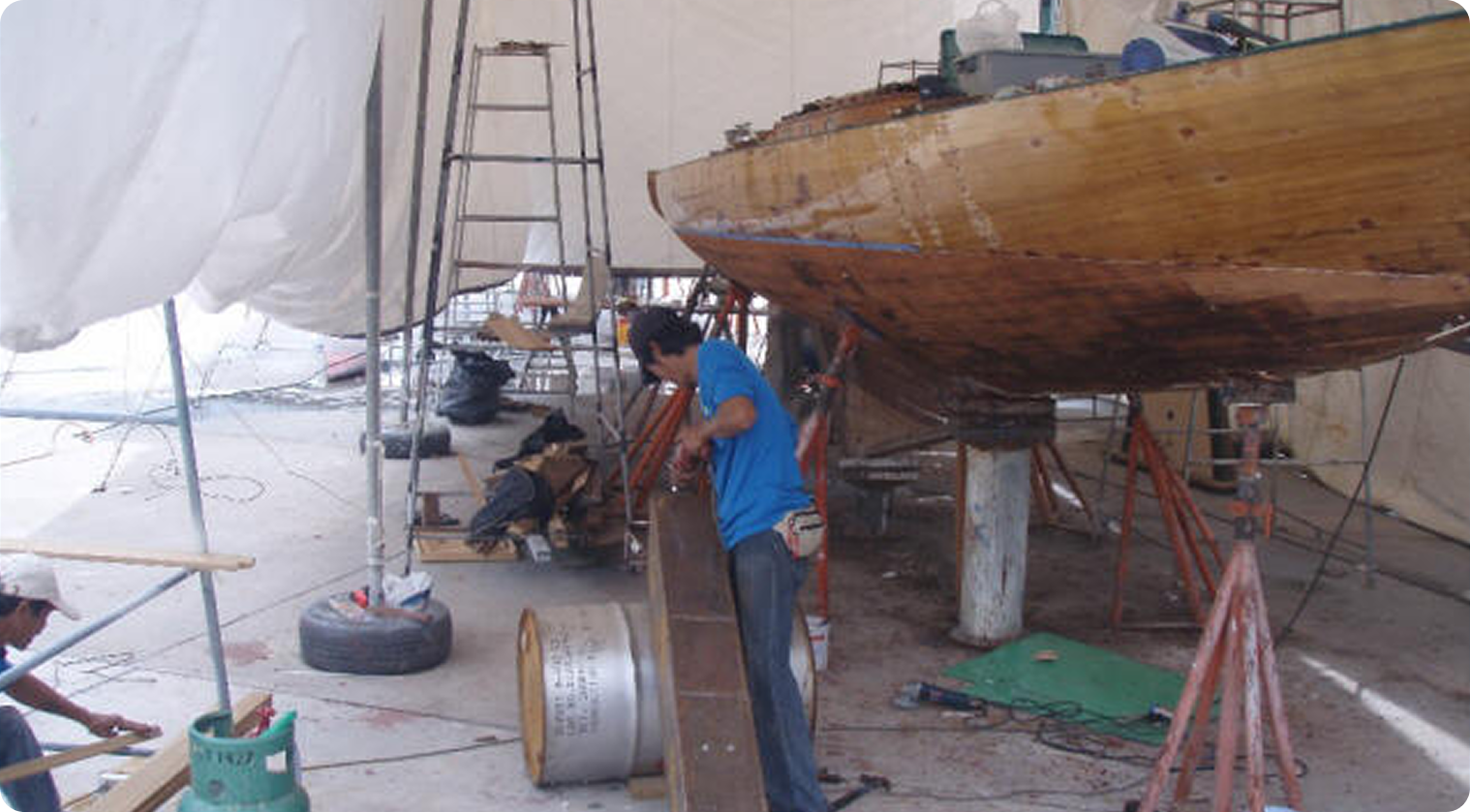 A man in a blue shirt works on the hull of a Classic Sailing Yacht in a large, covered workshop. Tools, ladders, and materials are scattered around, and another person is seated and working nearby.