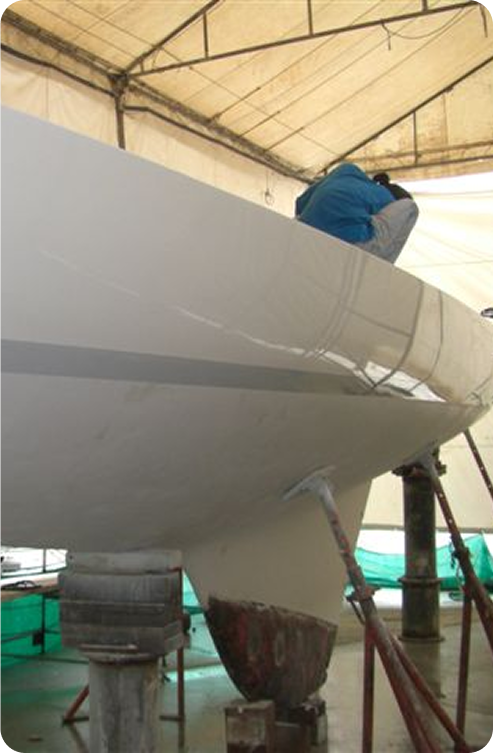 A person in a blue jacket works on the hull of a white Skerry Cruiser inside a covered workshop, with the boat elevated on supports.