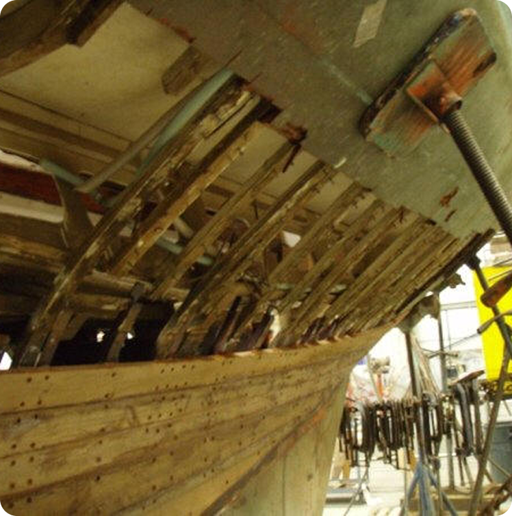 Partially restored wooden boat hull with exposed ribs and framework, showing ongoing repair work in a workshop setting. Tools and clamps are visible in the background, typical of classic sailing yachts restoration projects.