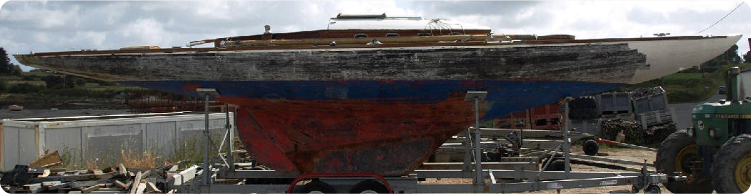 A weathered Skerry Cruiser with peeling paint and visible rust sits on a trailer in a boatyard, surrounded by grass, equipment, and a tractor under a cloudy sky.