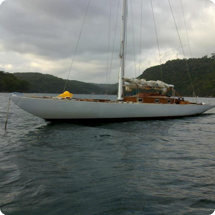 A classic white Skerry Cruiser with wooden details is anchored on calm water, surrounded by green, tree-covered hills under a cloudy sky.