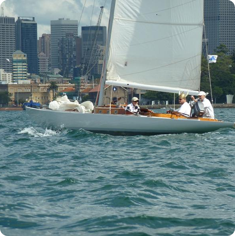 A classic sailing yacht with three people on board glides across choppy blue water, with tall city buildings and trees visible in the background under a partly cloudy sky.