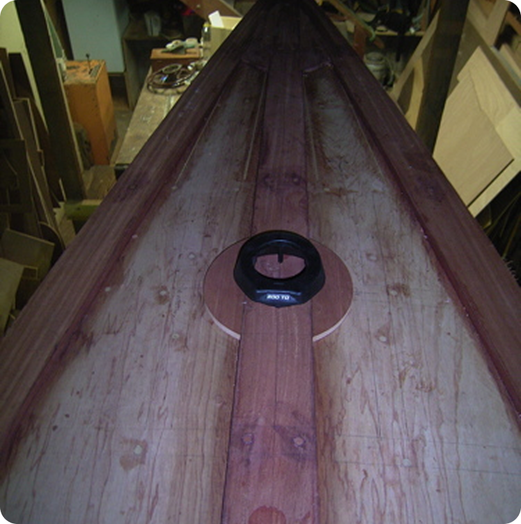 Close-up view of the wooden deck of a kayak under construction in a workshop, evoking the craftsmanship found in Skerry Cruisers, with a circular black hatch in the centre and various tools and materials in the background.