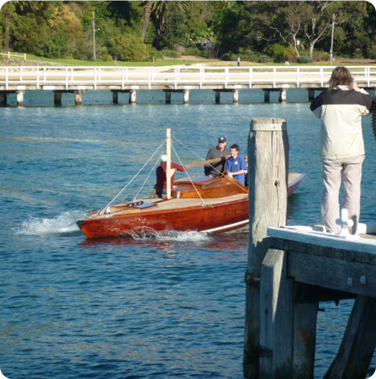 A classic sailing yacht with three people on board glides through the water near a quay, as someone onshore takes a photo. A white fence and green trees provide a scenic backdrop for this Square Metre Yachts moment.