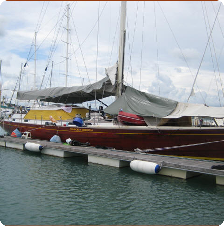 A red sailboat, reminiscent of Classic Sailing Yachts, with a grey tarpaulin covering its deck is moored at a marina. The boat is tied to a floating pontoon with several white and blue fenders along its side. The sky is cloudy.