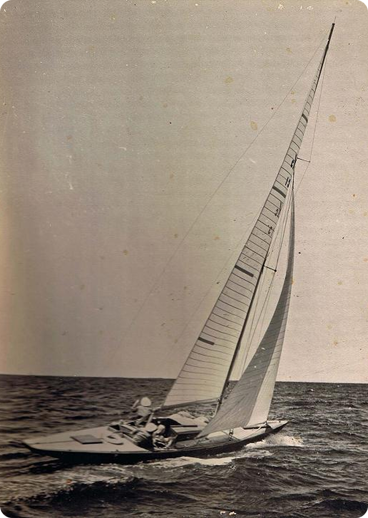 A vintage black-and-white photo of a Classic Sailing Yacht with a tall mast gliding on the sea, two people visible on deck, and waves breaking gently against the hull under an open sky.