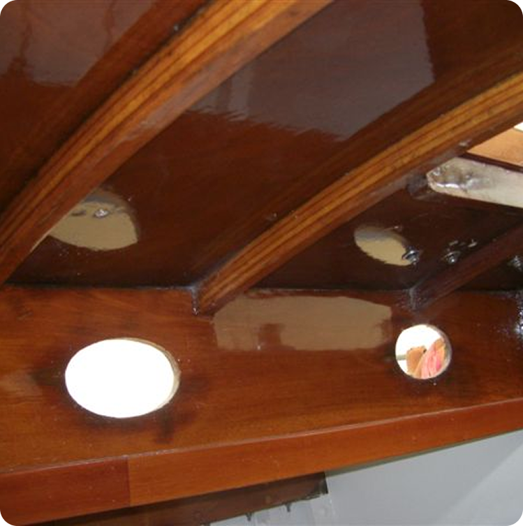 A close-up of a varnished wooden Skerry Cruiser interior shows curved beams and two round portholes, with light reflecting off the glossy wood surface.