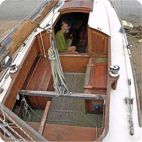 A person in a green shirt sits inside the wooden cabin of a small Skerry Cruiser sailboat, viewed from above. The boat is moored on dry land, with various ropes visible on the deck.