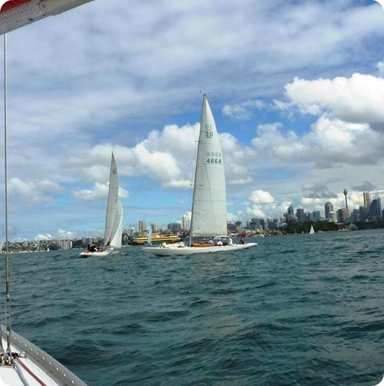 Several Square Metre Yachts glide across a body of water on a partly cloudy day, with a city skyline and tall buildings in the background. The scene is peaceful, featuring classic sailing yachts amid rippling waves and blue skies.