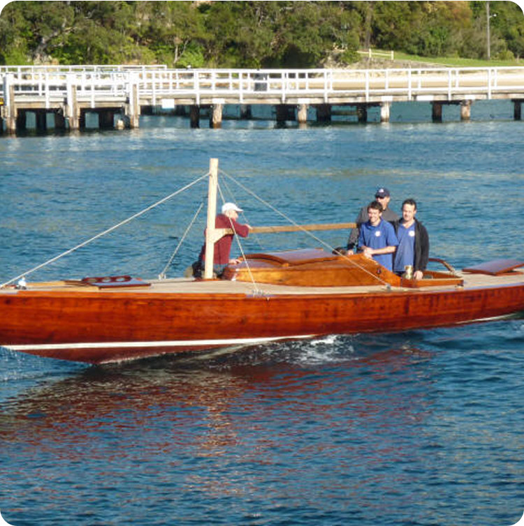 A sleek, polished wooden Square Metre Yacht with four people aboard moves across blue water near a white pier and tree-lined shore on a sunny day.
