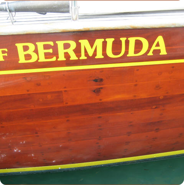 The side of a wooden boat, likely inspired by Classic Sailing Yachts, with the word BERMUDA painted in large yellow letters on a reddish-brown surface above a yellow stripe near the waterline.
