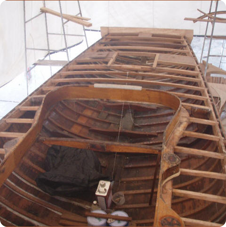 A wooden Classic Sailing Yacht under restoration, its deck partially removed to reveal the framework and ribs. Scaffolding surrounds the vessel, with various tools and materials scattered inside the exposed hull.