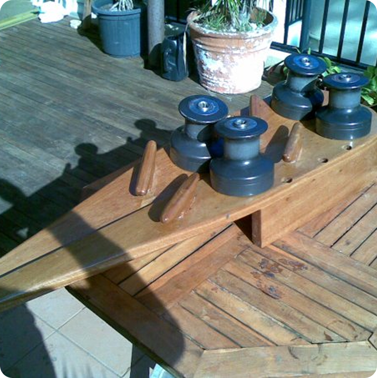 A wooden wine rack shaped like the bow of a Square Metre Yacht sits on a wooden table outdoors, with four wine bottle holders resembling winches and potted plants in the background.