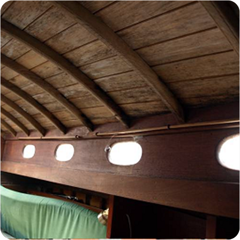 Interior view of a wooden Skerry Cruisers boat cabin roof with curved beams and small oval windows letting in light; part of a covered green cushion is visible below the windows.