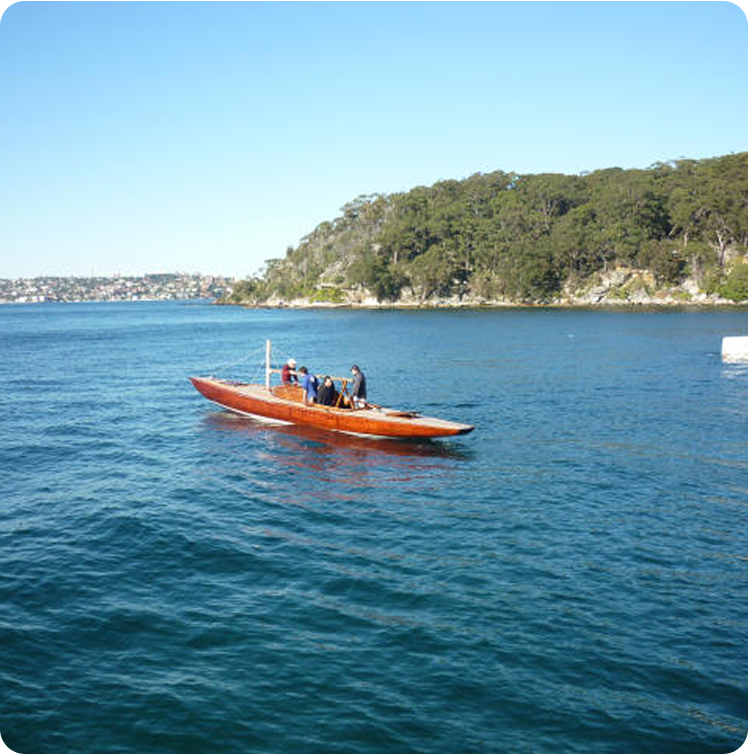 A small wooden boat, reminiscent of classic sailing yachts, floats on calm blue water near a tree-covered shoreline under a clear sky.