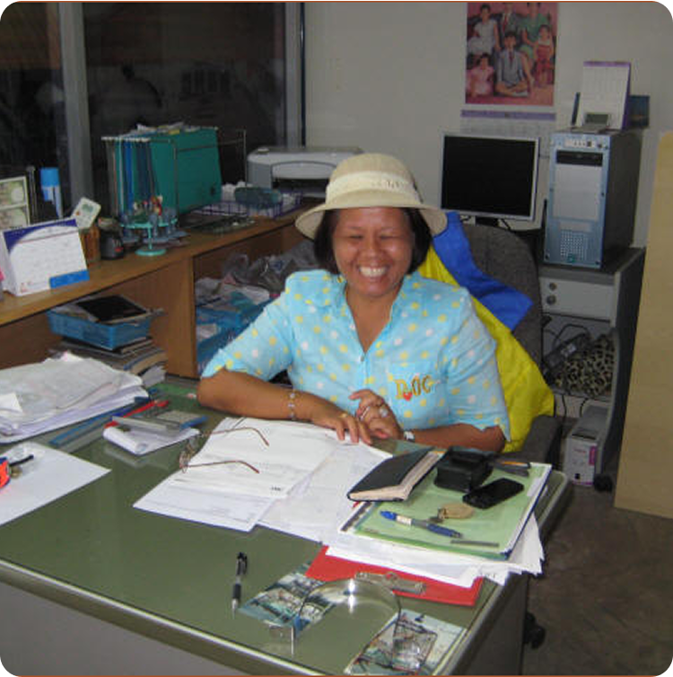 A woman wearing a hat and a blue polka-dot shirt sits at a cluttered desk, smiling widely. Among the office supplies are magazines featuring Classic Sailing Yachts, adding a nautical touch to the busy workspace.