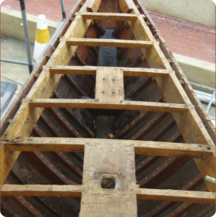 A wooden boat frame with exposed ribs and crossbeams, possibly a Schärenkreuzer or another type of Classic Sailing Yacht, is under construction or restoration outdoors on a paved surface with a traffic cone nearby.