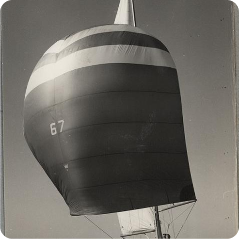A black-and-white photo of a Schärenkreuzer sailboat with the number 67 on its large, billowing striped sail. The sail fills most of the image, and the sky in the background is clear.